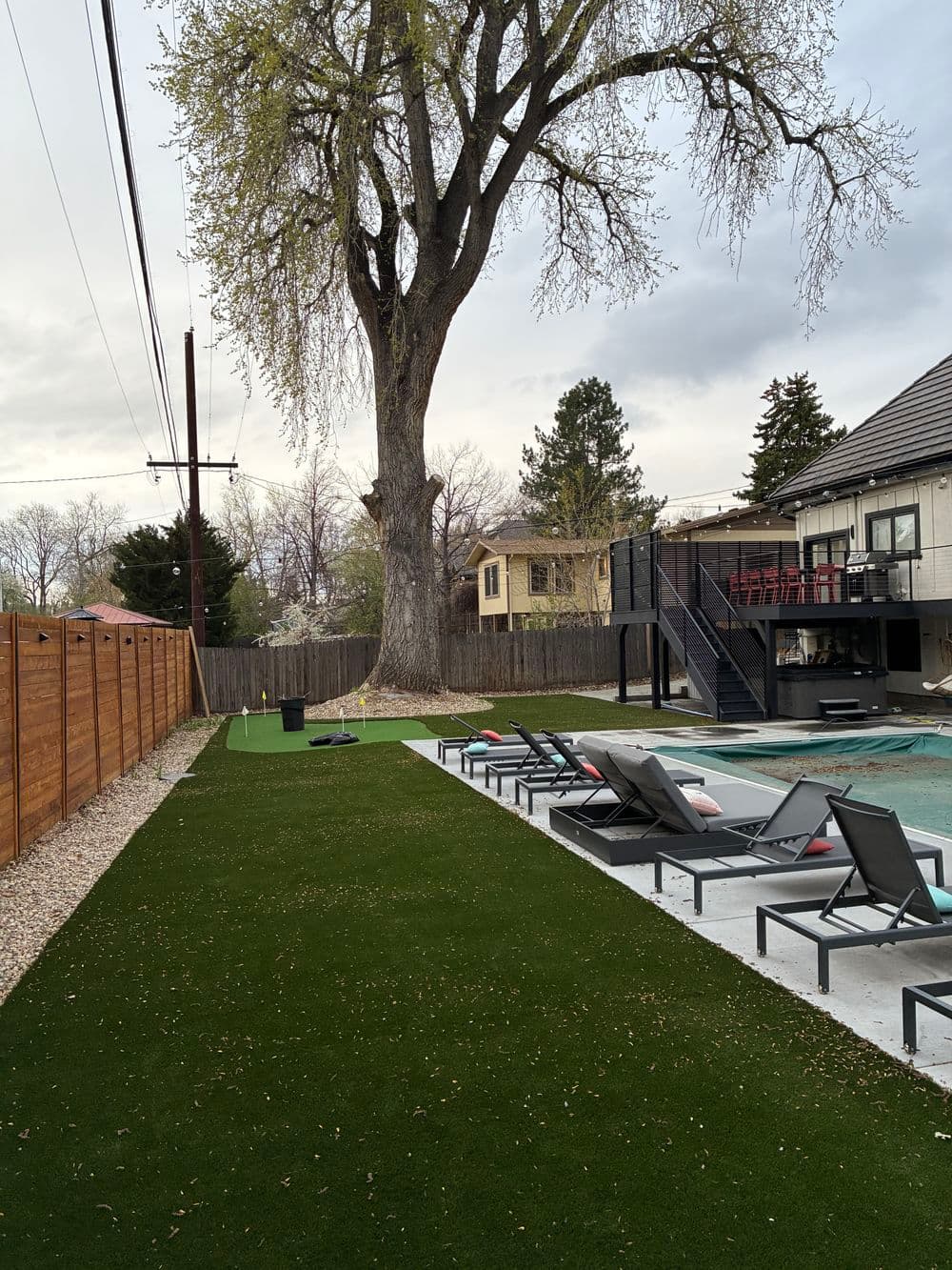 Backyard pool area with artificial grass, lounge chairs, and a large tree in view.